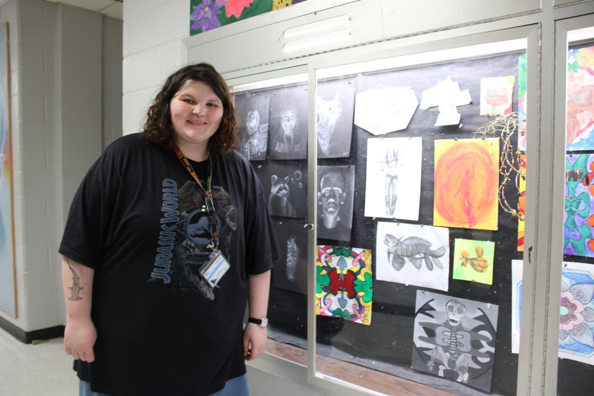 In the art hallway, Ariel Harman stands with some of her displayed work which includes an illustration of a prehistoric shark.