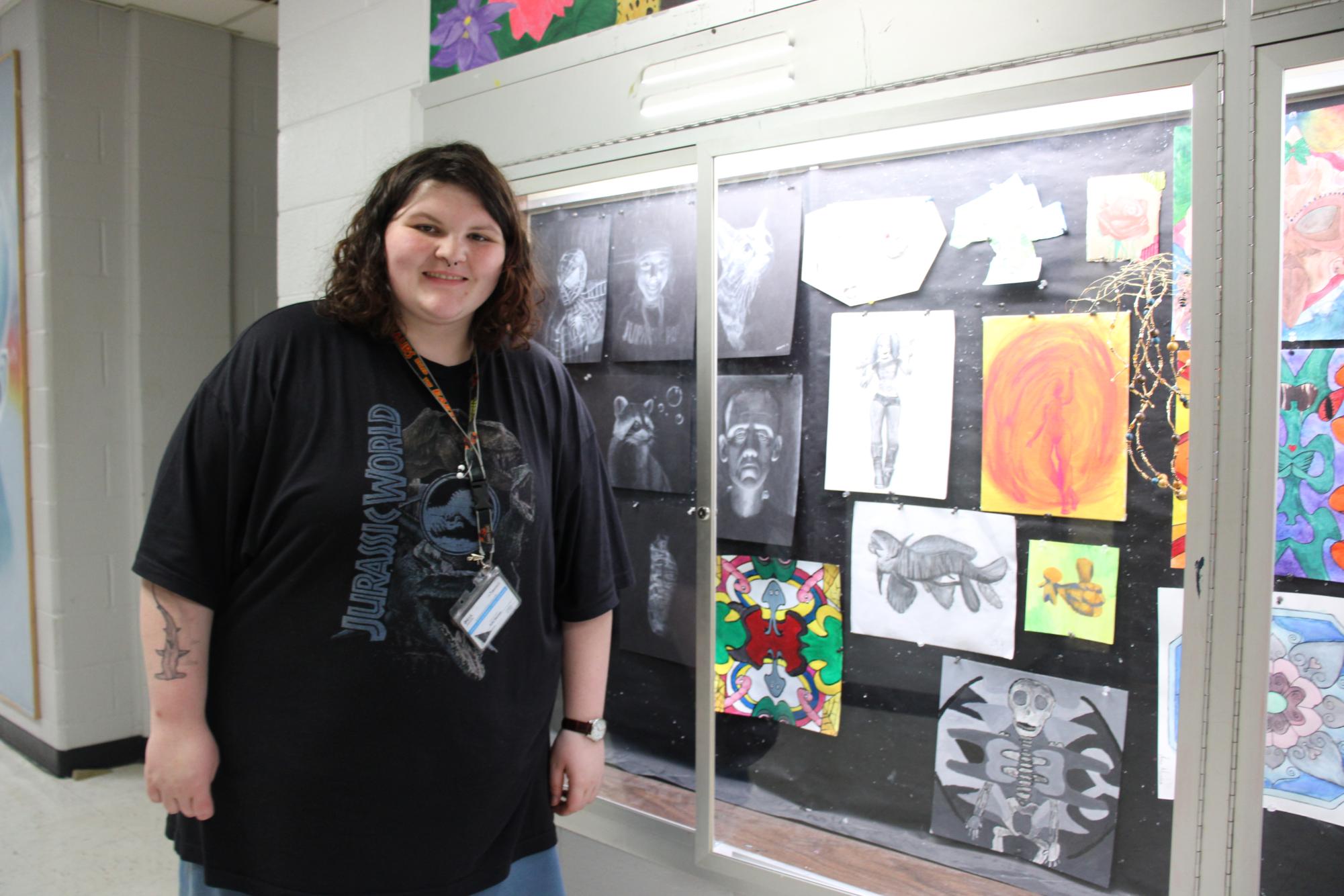 In the art hallway, Ariel Harman stands with some of her displayed work which includes an illustration of a prehistoric shark.